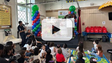 A man reads a big book to children on a mat with First 5 San Bernardino's mascot Potter the Otter and a video play button overlay indicating a video.