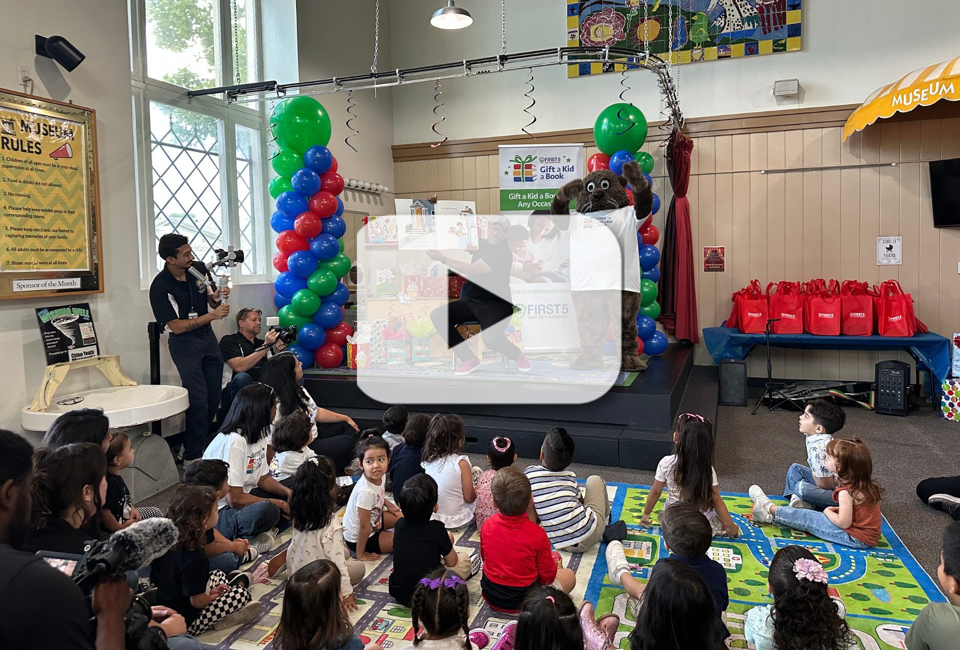 A man reads a big book to children on a mat with First 5 San Bernardino's mascot Potter the Otter and a video play button overlay indicating a video.