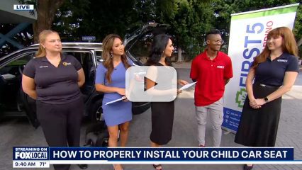 Five people, including news reporters, stand near a car during a live Fox TV broadcast highlighting child car seat safety.