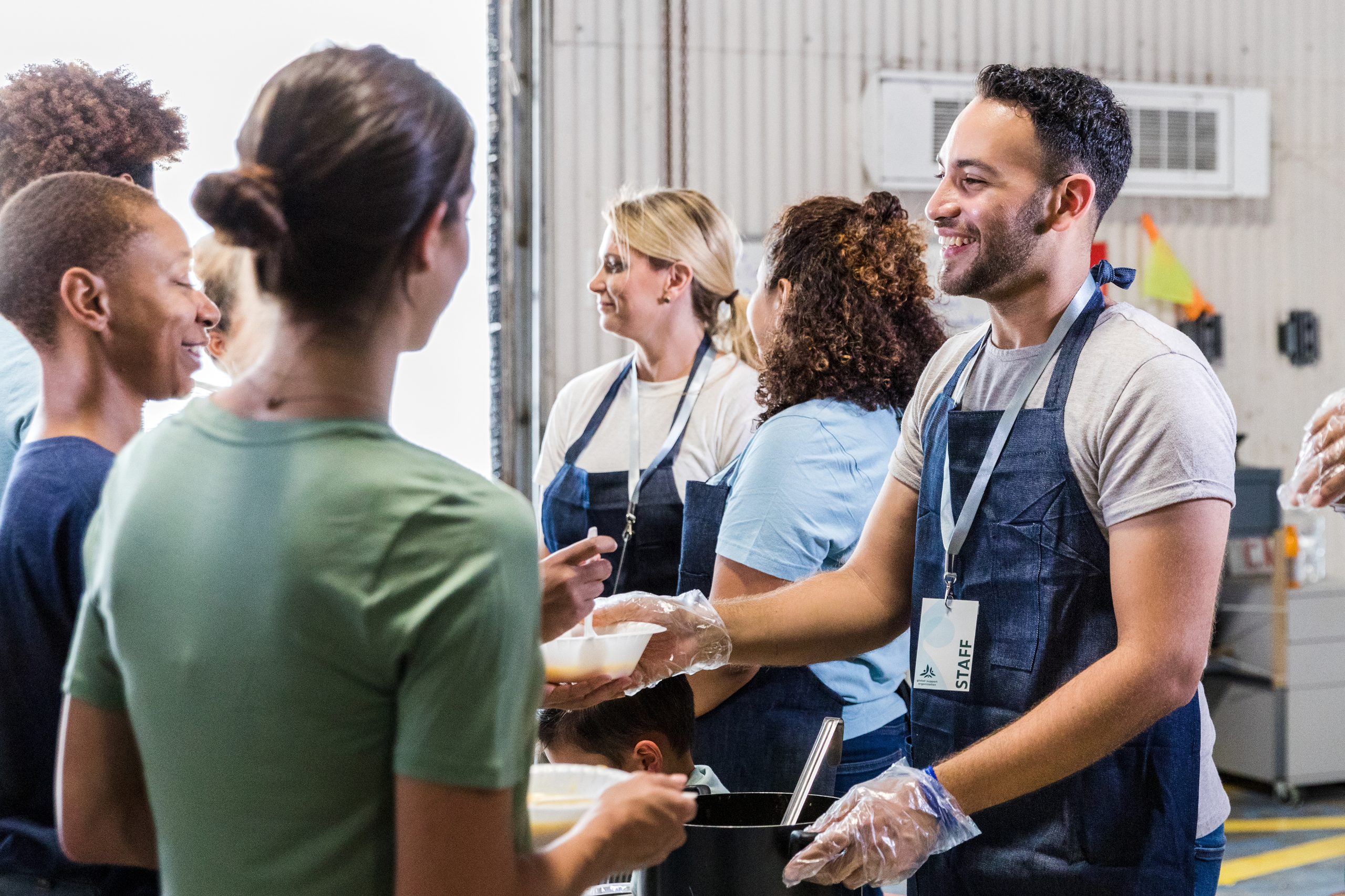 A staff member wearing an apron and gloves serves food to community members while other volunteers assist at a food distribution site.