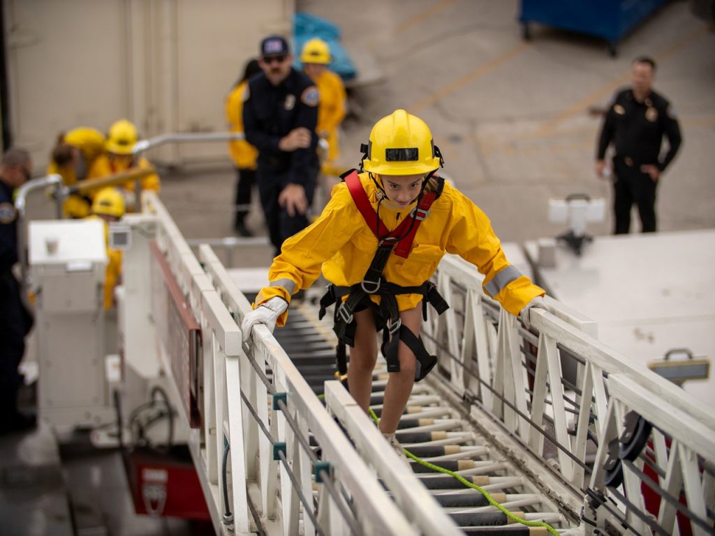 Girl Empowerment Camp participant in a yellow helmet and safety harness climbs an aerial ladder while firefighters look on.