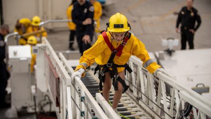 Girl Empowerment Camp participant in a yellow helmet and safety harness climbs an aerial ladder while firefighters look on.