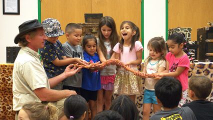 A group of excited children and a man hold a large snake during an educational event. The atmosphere is lively, with kids showing curiosity and joy.