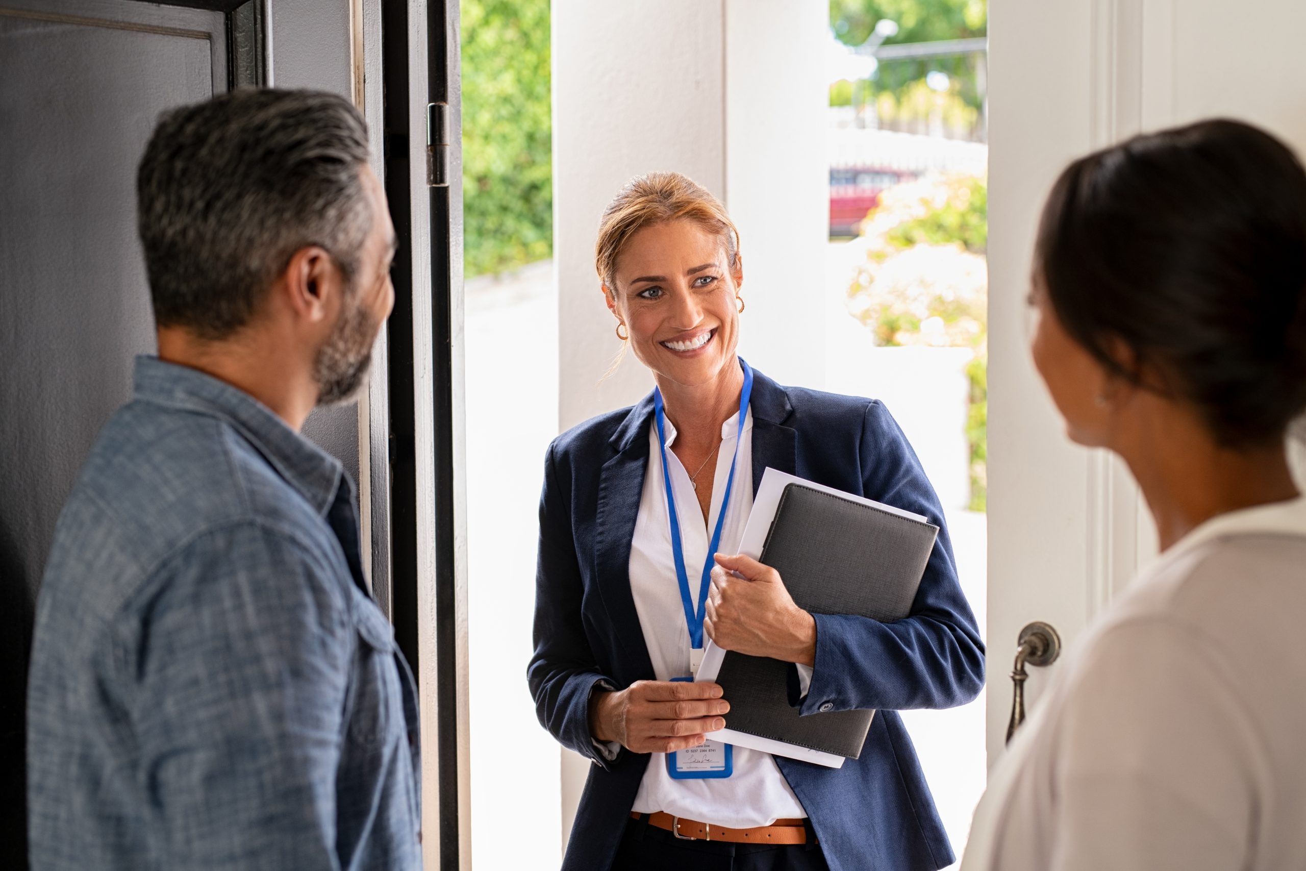 A female caseworker stands at a home's front door, speaking with two residents while holding a folder.