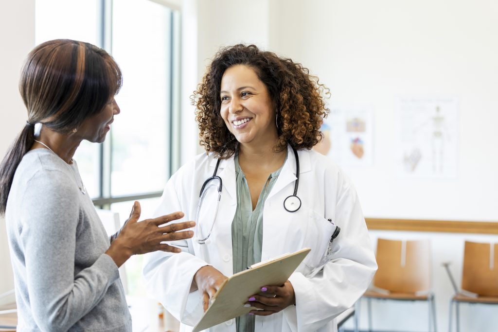A female doctor holding a clipboard smiles at a female patient in a clinic setting.