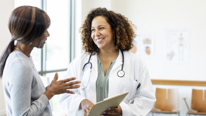 A female doctor holding a clipboard smiles at a female patient in a clinic setting.