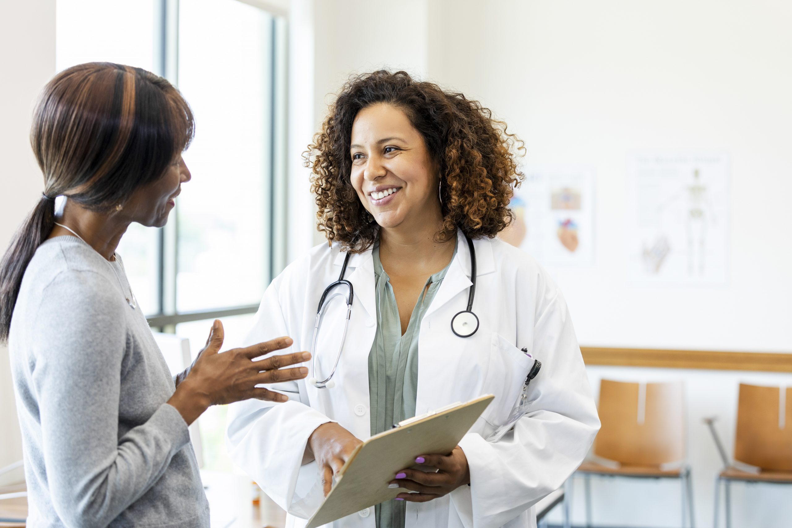 A female doctor holding a clipboard smiles at a female patient in a clinic setting.