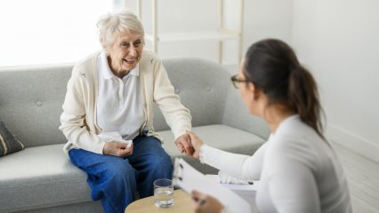 An older adult sits on a couch holding a tisue and smiling while holding a professional woman’s woman.