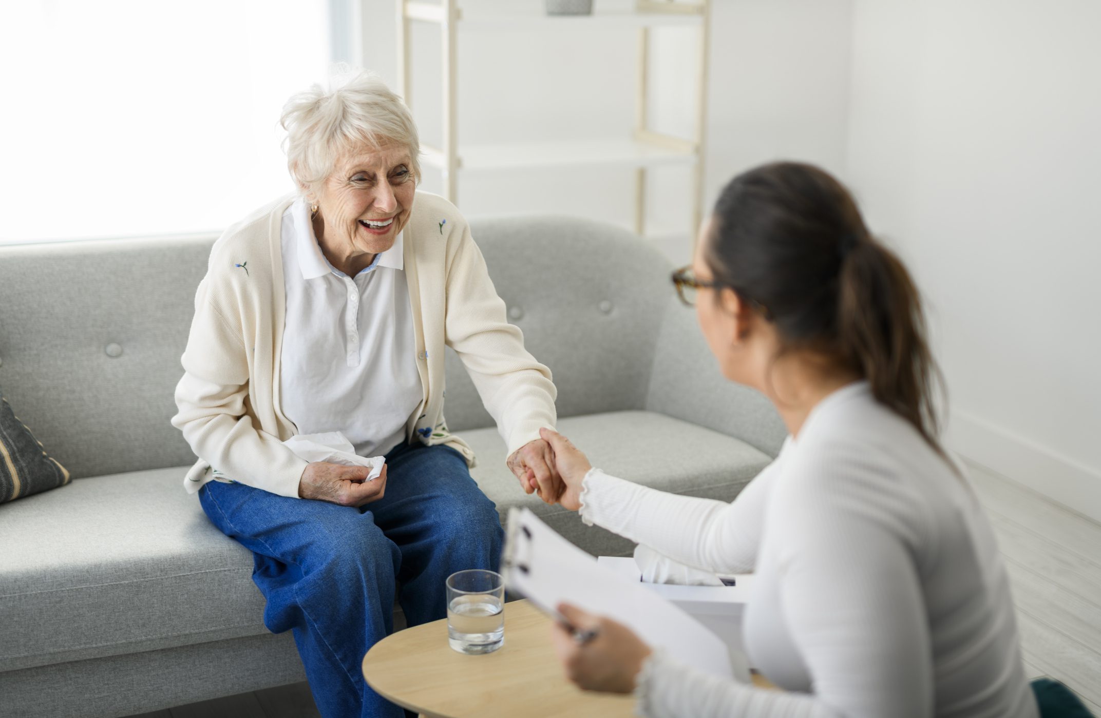 An older adult sits on a couch holding a tisue and smiling while holding a professional woman’s woman.