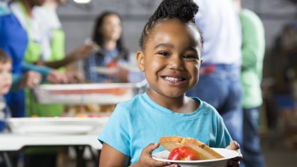 A young girl smiles, wearing a blue T-shirt and holding a bowl with an apple and a slice of bread in it.