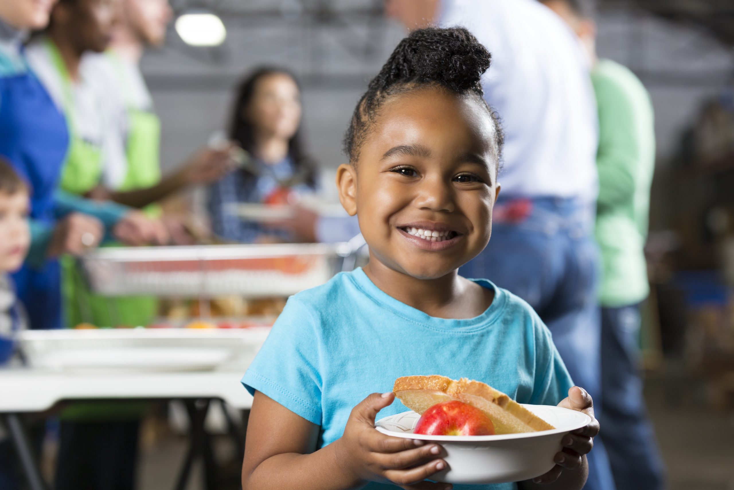 A young girl smiles, wearing a blue T-shirt and holding a bowl with an apple and a slice of bread in it.