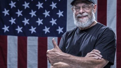 Person showing a thumbs up in front of an American flag, wearing glasses and a black cap that says Vietnam Veteran.