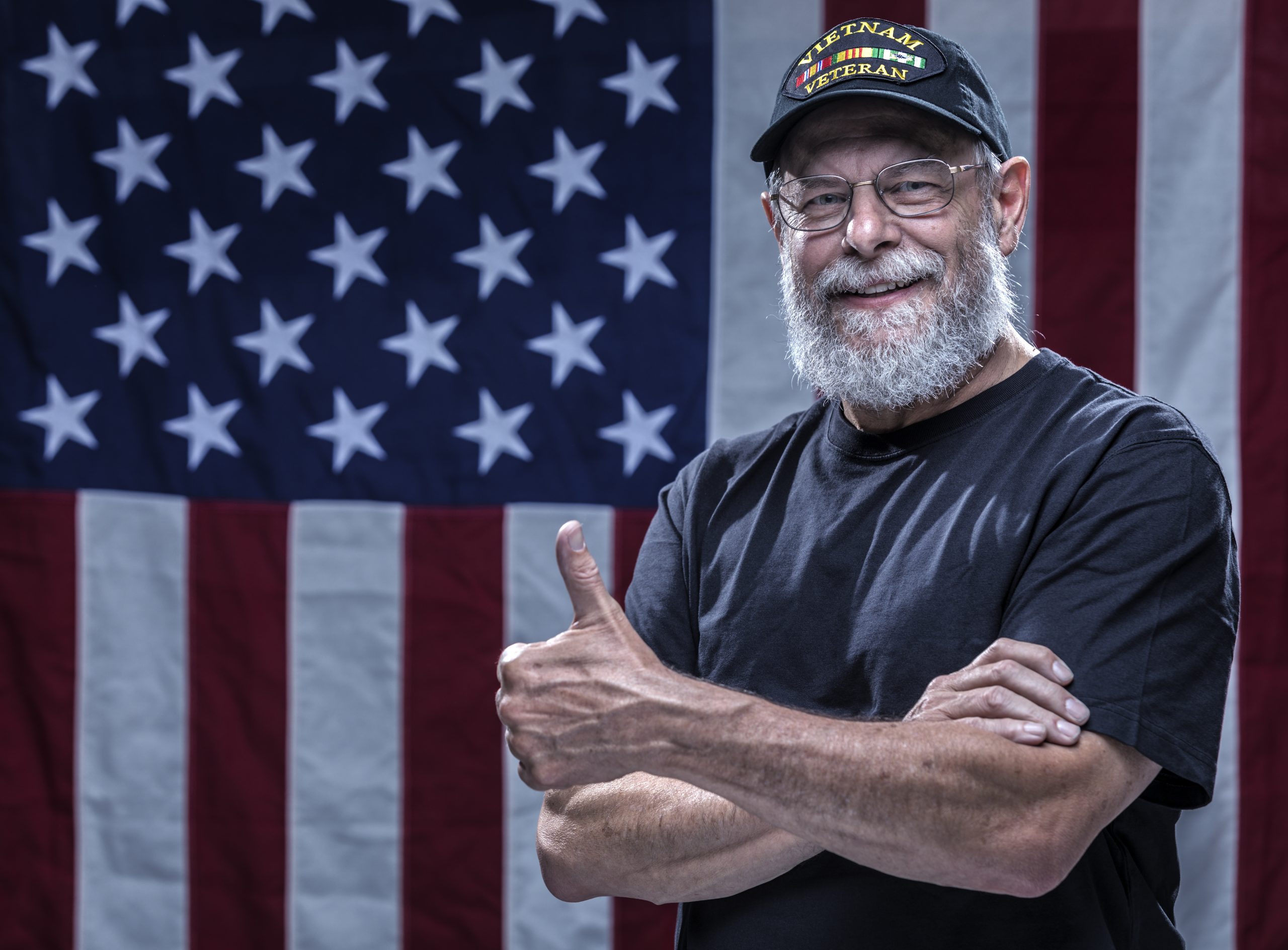 Person showing a thumbs up in front of an American flag, wearing glasses and a black cap that says Vietnam Veteran.