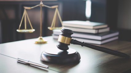 A wood gavel rests on a desk with a stack of books and the scales of justice.