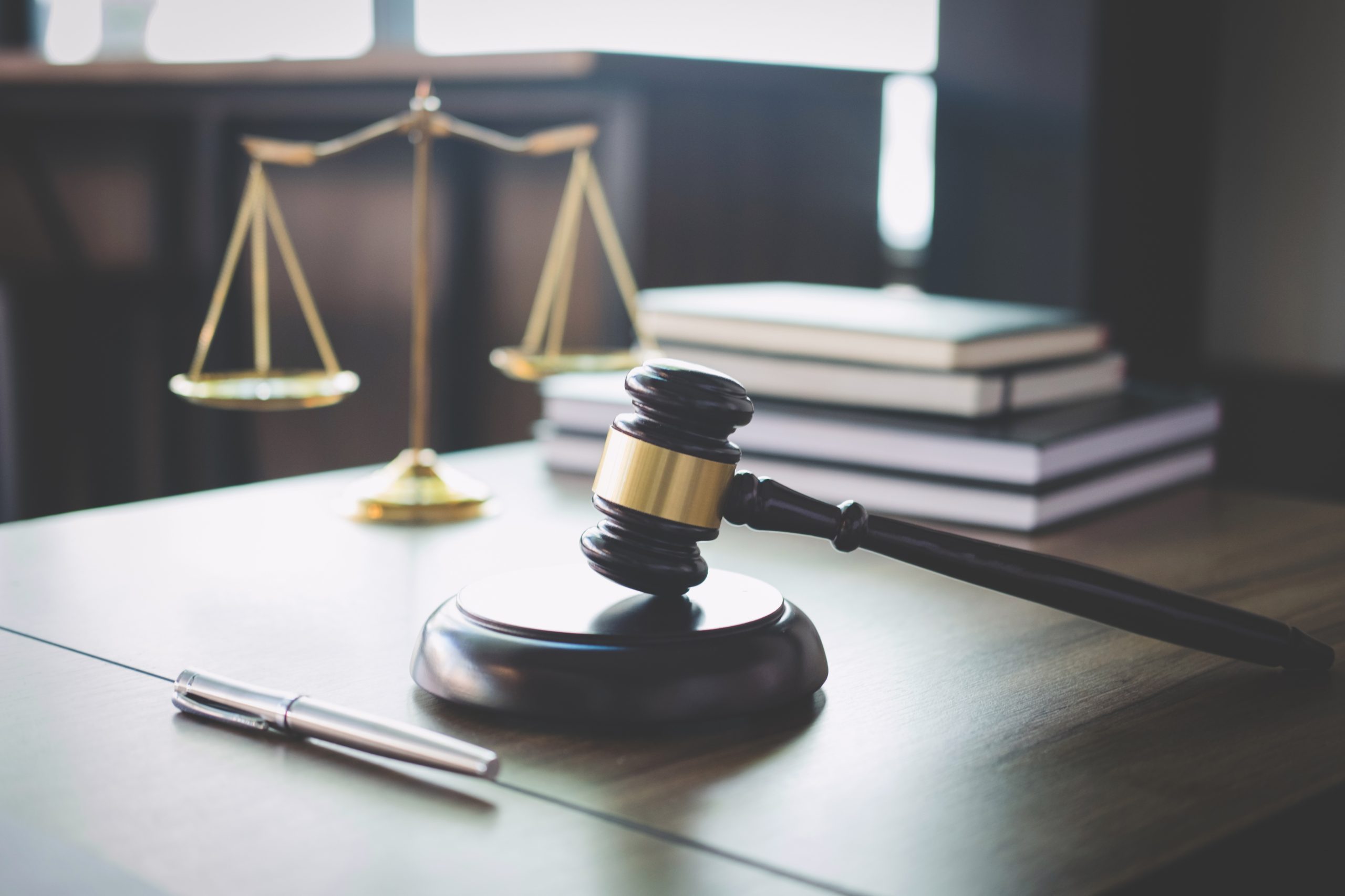 A wood gavel rests on a desk with a stack of books and the scales of justice.