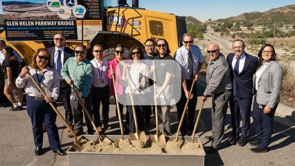 Public Works staff smiling in celebration of a groundbreaking event, lifting shovels of sand in front of a bright yellow construction machine on a sunny day.