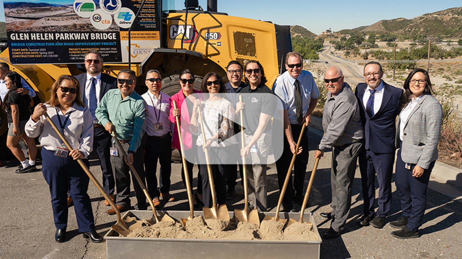 Public Works staff smiling in celebration of a groundbreaking event, lifting shovels of sand in front of a bright yellow construction machine on a sunny day.