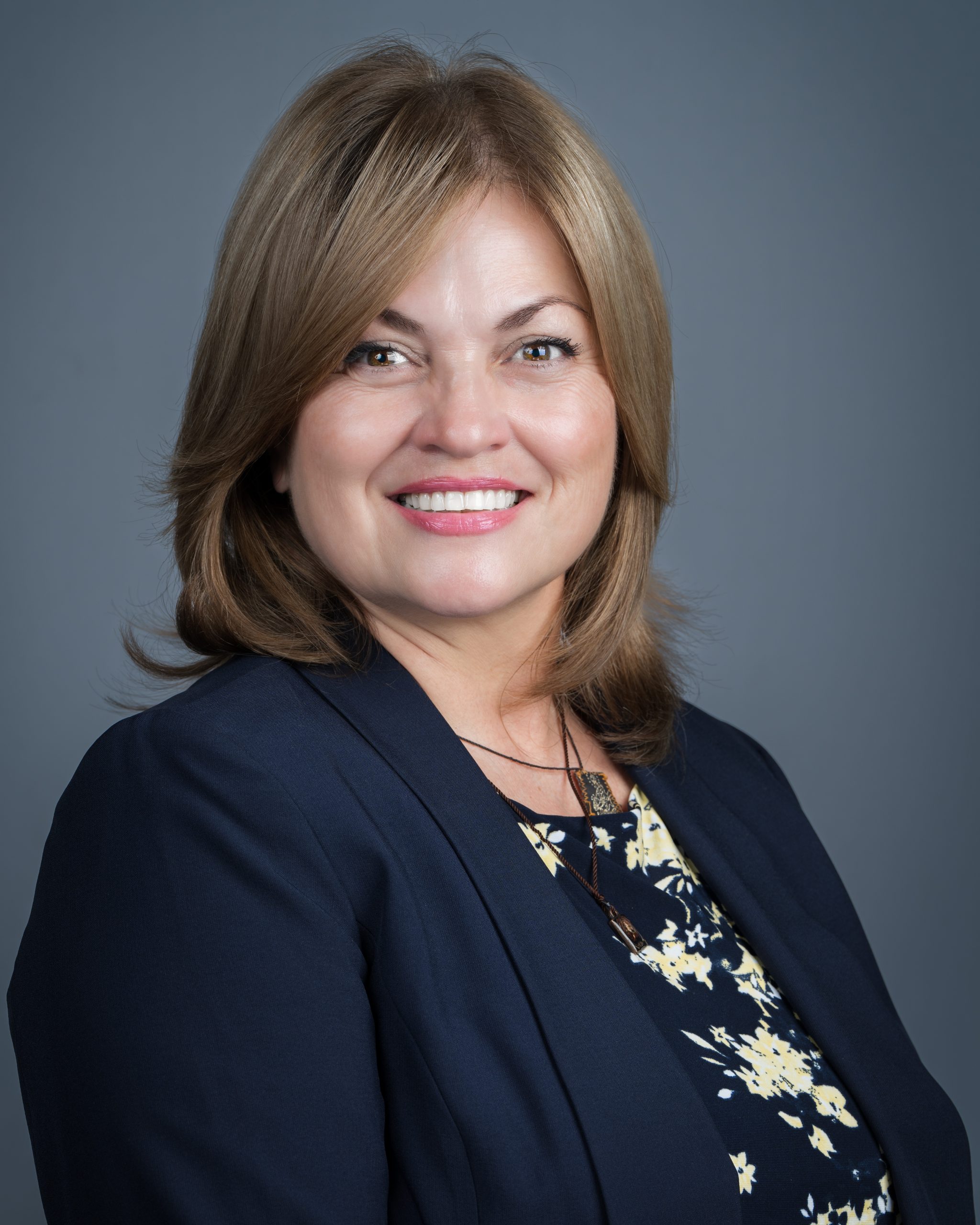 Headshot of a woman with shoulder-length brown hair smiling, wearing a navy blazer and patterned blouse on a gray background.