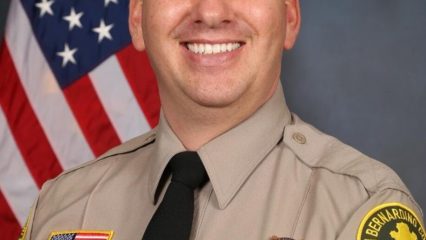 A person in San Bernardino County Sheriff's uniform is smiling. The uniform displays badges and an American flag patch. The background features a U.S. flag.