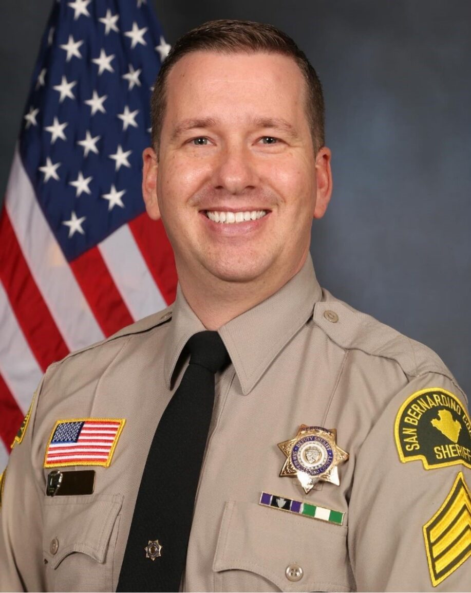 A person in San Bernardino County Sheriff's uniform is smiling. The uniform displays badges and an American flag patch. The background features a U.S. flag.