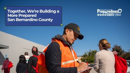 A man in an orange safety vest writes on a clipboard as people with red backpacks line up outdoors. Text reads: “Together, We’re Building a More Prepared San Bernardino County” and “Preparedness Starts Here.”