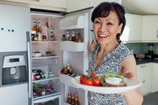 A woman stands by an open refrigerator holding a plate of nutritious food.