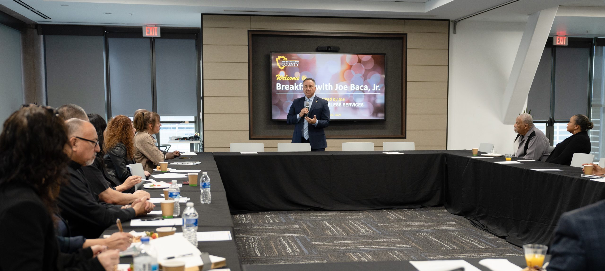 Supervisor Joe Baca, Jr. stands at the front of a conference room, welcoming attendees to his homelessness roundtable.