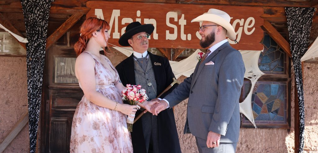 A woman and man stand facing each other on the Main Street Stage at Calico Ghost Town with a man dressed in 1800 period clothing standing in between looking at the man.