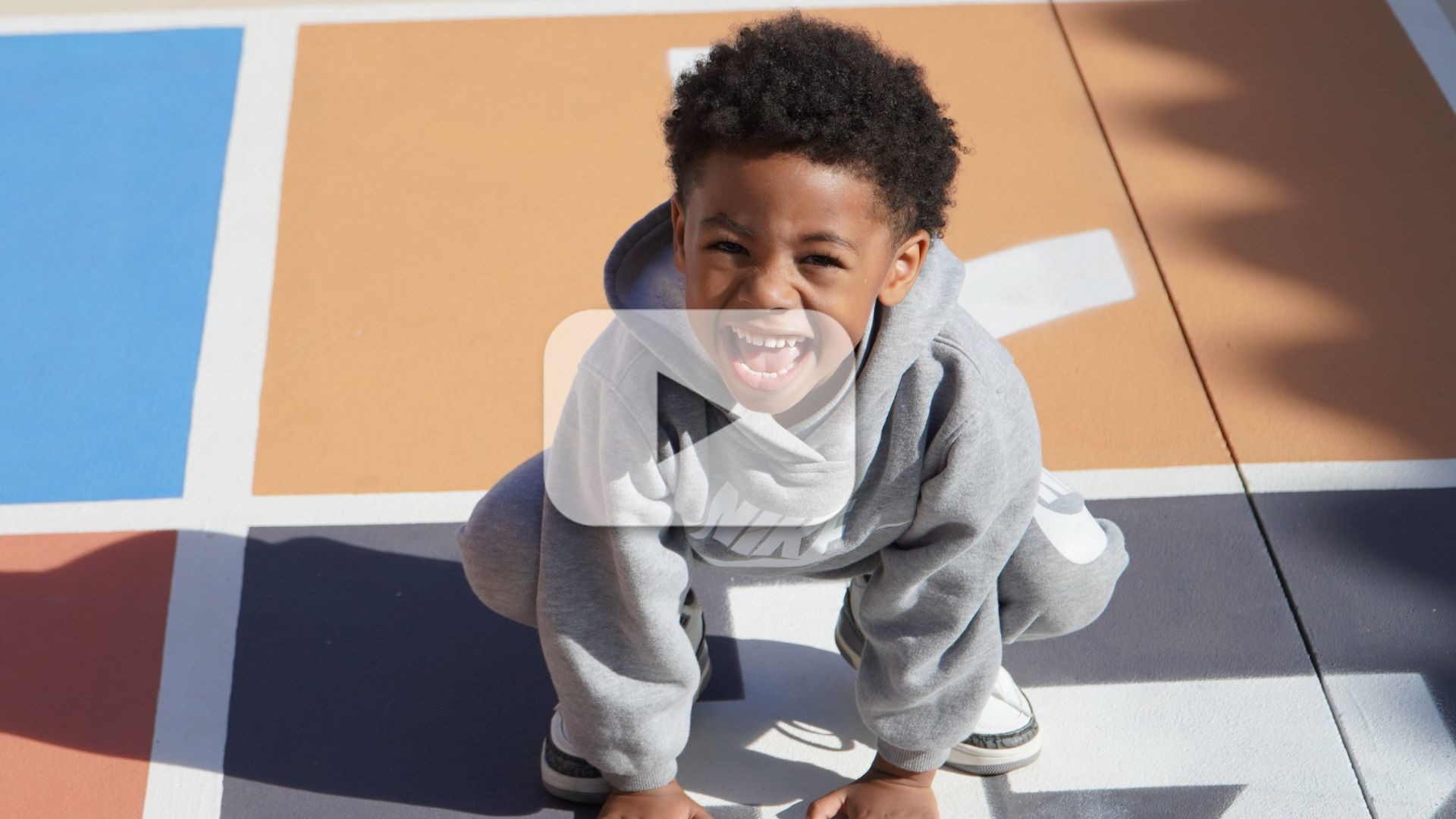 Smiling child crouches on a colorful playground surface, looking up at the camera in bright sunlight, with a play button overlay indicating a video.