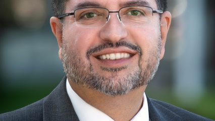 Carlos Prieto, a man wearing a dark suit, white shirt, and blue patterned tie, with glasses, and a trimmed beard, standing outdoors with a blurred background of greenery and building.