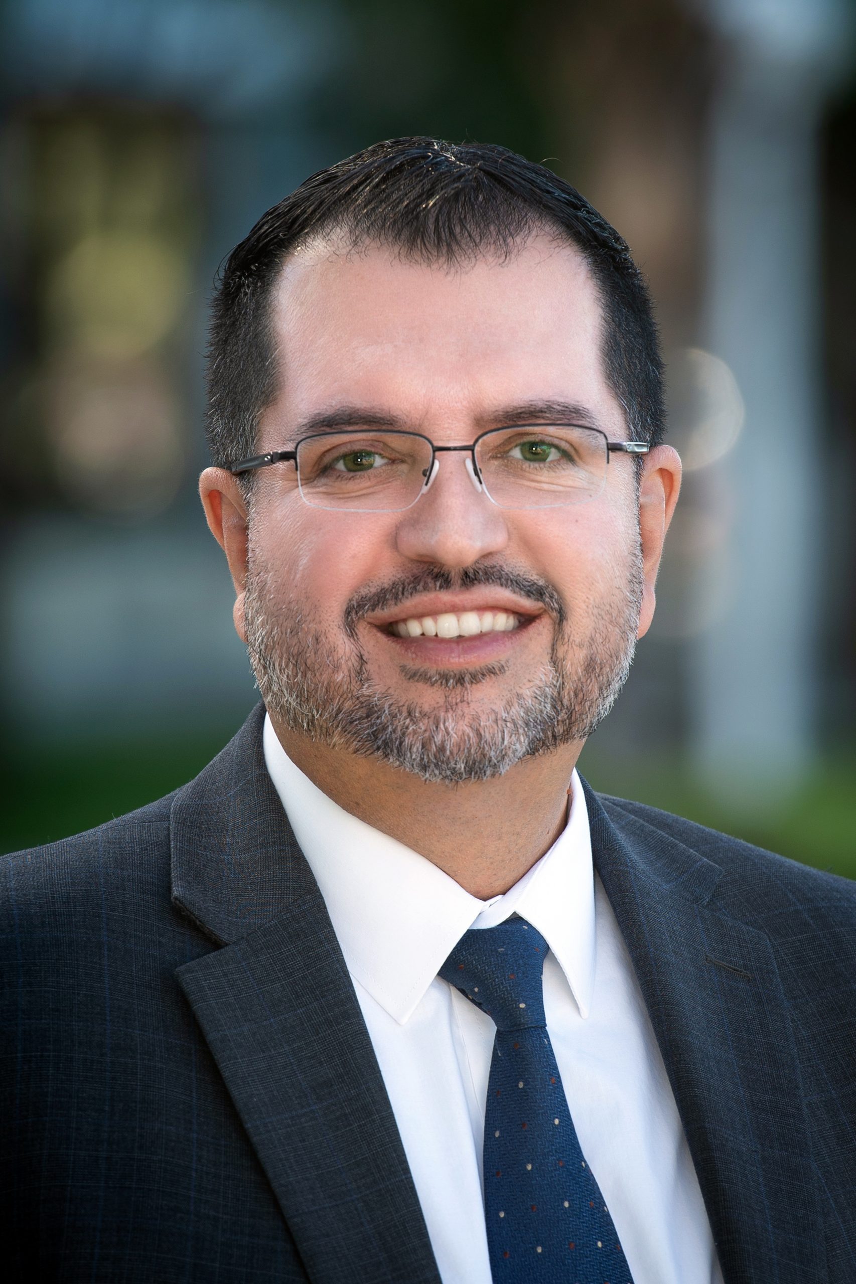 Carlos Prieto, a man wearing a dark suit, white shirt, and blue patterned tie, with glasses, and a trimmed beard, standing outdoors with a blurred background of greenery and building.