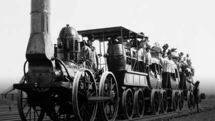 A black-and-white photograph of an early steam-powered locomotive pulling several open carriages with passengers in traditional 19th-century clothing. The train's industrial design features a riveted exterior, large metal wheels and a chimney-like smokestack.