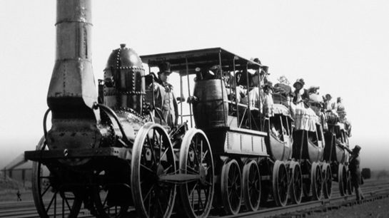 A black-and-white photograph of an early steam-powered locomotive pulling several open carriages with passengers in traditional 19th-century clothing. The train's industrial design features a riveted exterior, large metal wheels and a chimney-like smokestack.