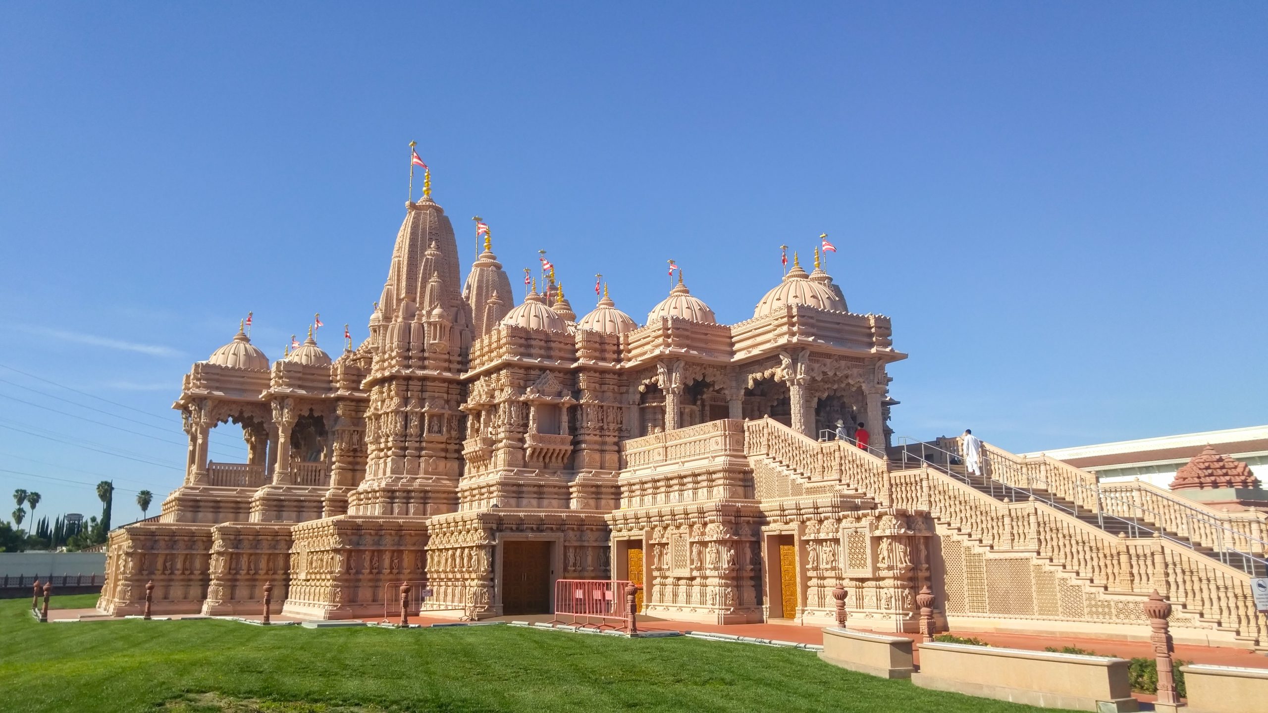 Exterior view of the BAPS Shri Swaminarayan Mandir, displaying intricate architecture with multiple domes, crafted in pink sandstone, under a clear blue sky.