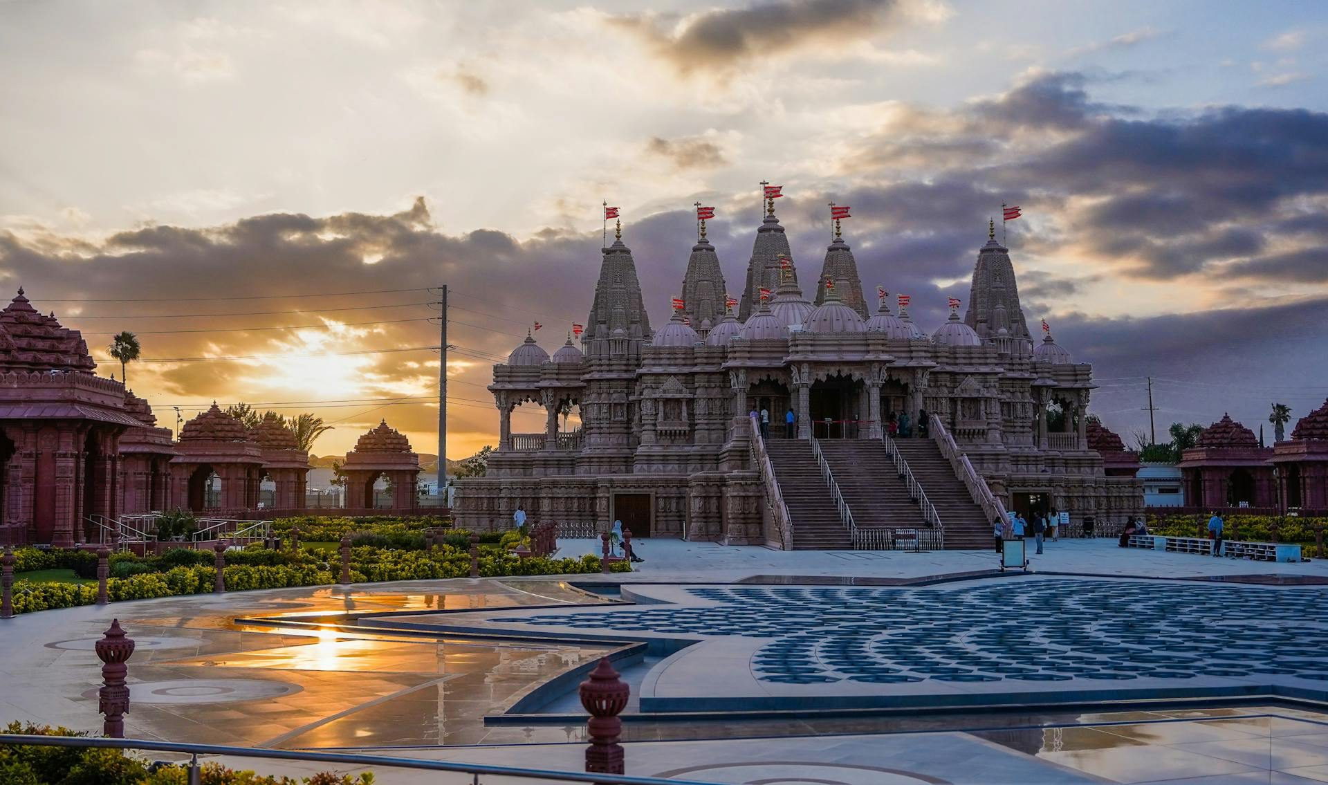 Sunset view of the BAPS Hindu Temple in Chino Hills, featuring the grand temple with intricate architecture against a vibrant sky, and several visitors walking around the spacious temple grounds.