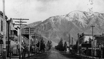 Black and white photo of a main street with businesses aligned on each side and mountains in the background.