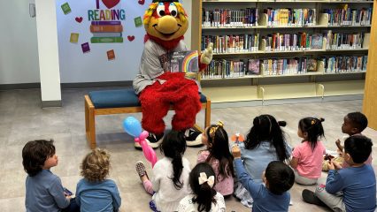 Mascot with a colorful outfit and cap reads a book to a group of young children seated on the floor in a library.