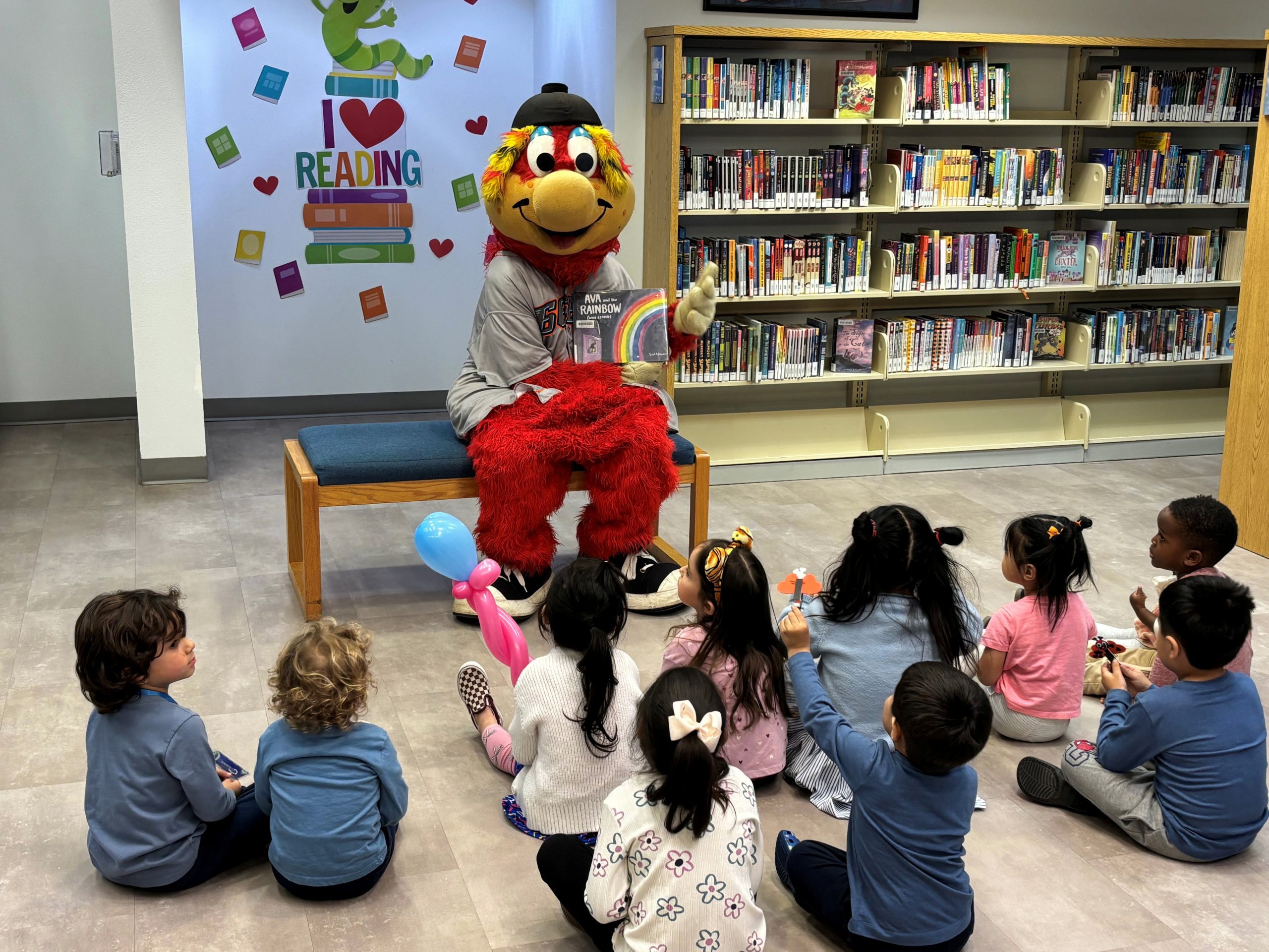 Mascot with a colorful outfit and cap reads a book to a group of young children seated on the floor in a library.
