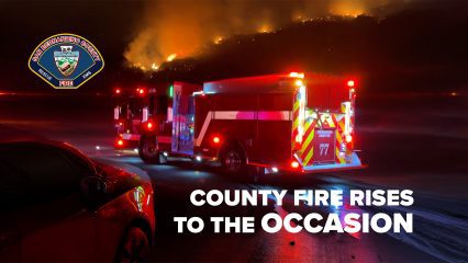 A nighttime scene shows a fire truck positioned on a road, responding to a significant fire on a hillside. Also visible is the County Fire logo and text that reads County Fire Rises To The Occasion.