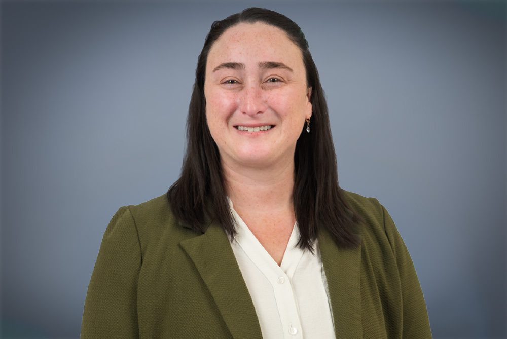 A headshot of a smiling woman wearing an olive blazer and white blouse against a soft gray background.