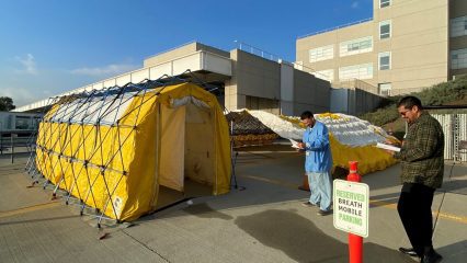 Two people with clipboards observe the set up of a medical modular shelter.