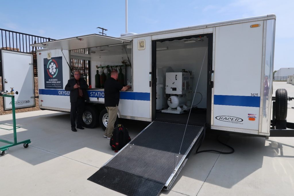 Two individuals stand beside an open trailer labeled "Oxygen Station," equipped with medical supplies and setup for emergency services.