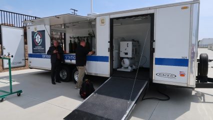Two individuals stand beside an open trailer labeled "Oxygen Station," equipped with medical supplies and setup for emergency services.