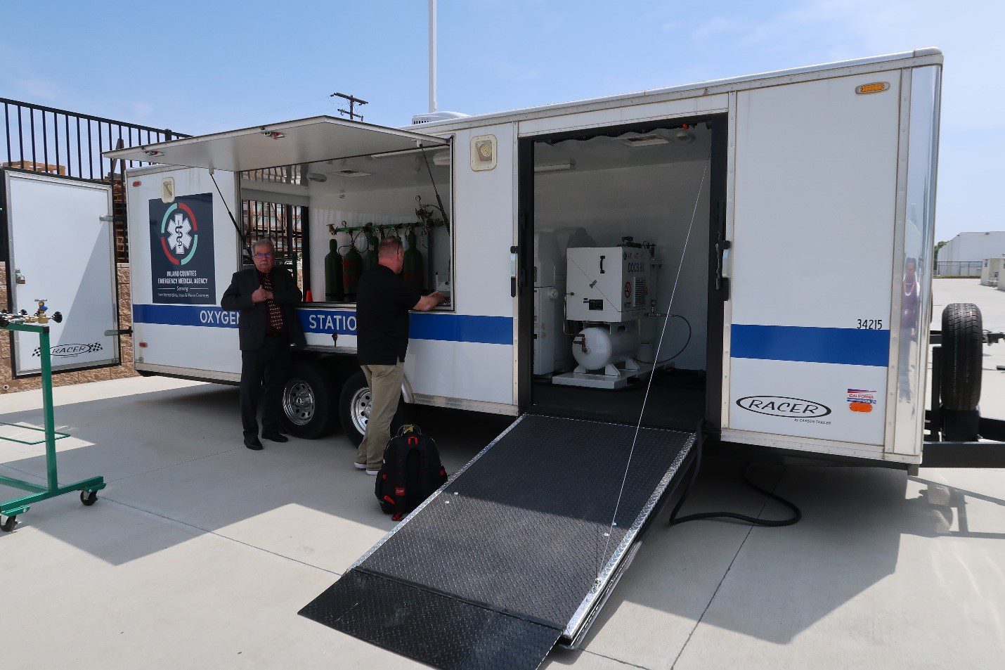 Two individuals stand beside an open trailer labeled "Oxygen Station," equipped with medical supplies and setup for emergency services.