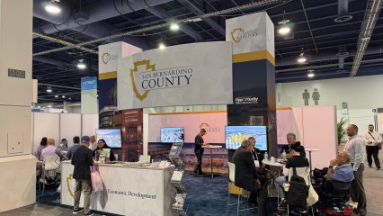 San Bernardino County Economic Development booth at a trade show with signage, displays, brochures, seating, and attendees networking.