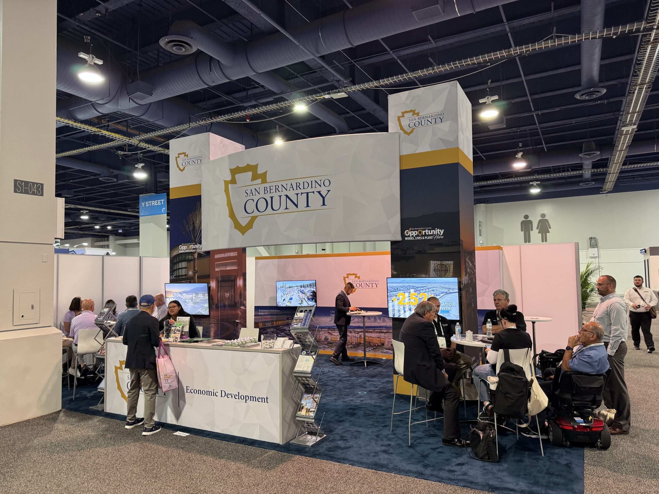 San Bernardino County Economic Development booth at a trade show with signage, displays, brochures, seating, and attendees networking.
