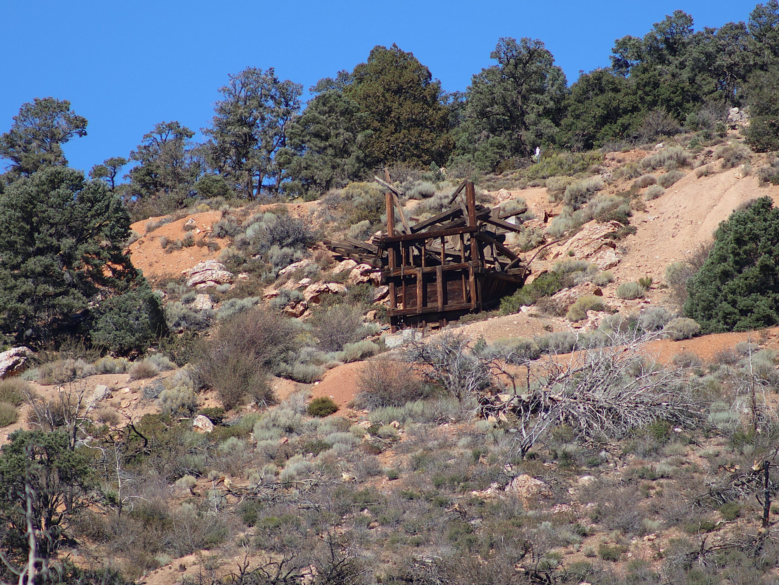 A wooden structure is situated on a rocky hillside amid sparse vegetation and bushes. The landscape features scattered trees and dry brush under a clear blue sky.