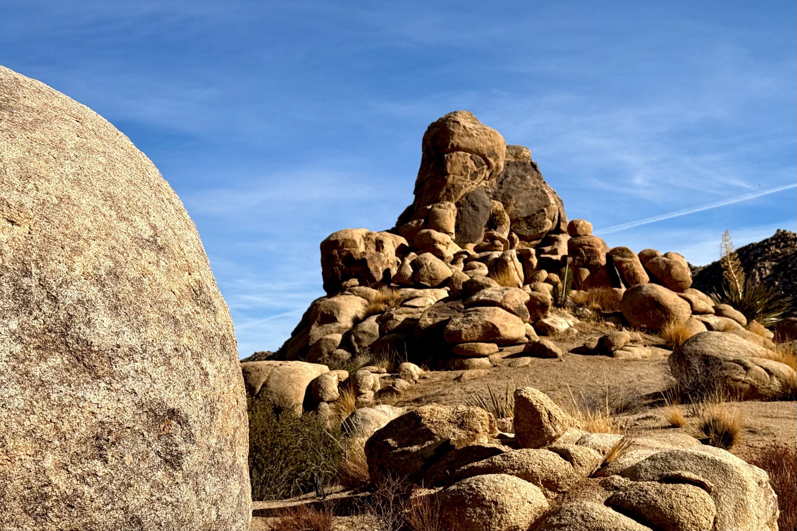 A landscape featuring a collection of large, weathered boulders under a clear blue sky. The foreground includes a prominent rounded boulder. Sparse vegetation is visible among the rocks.