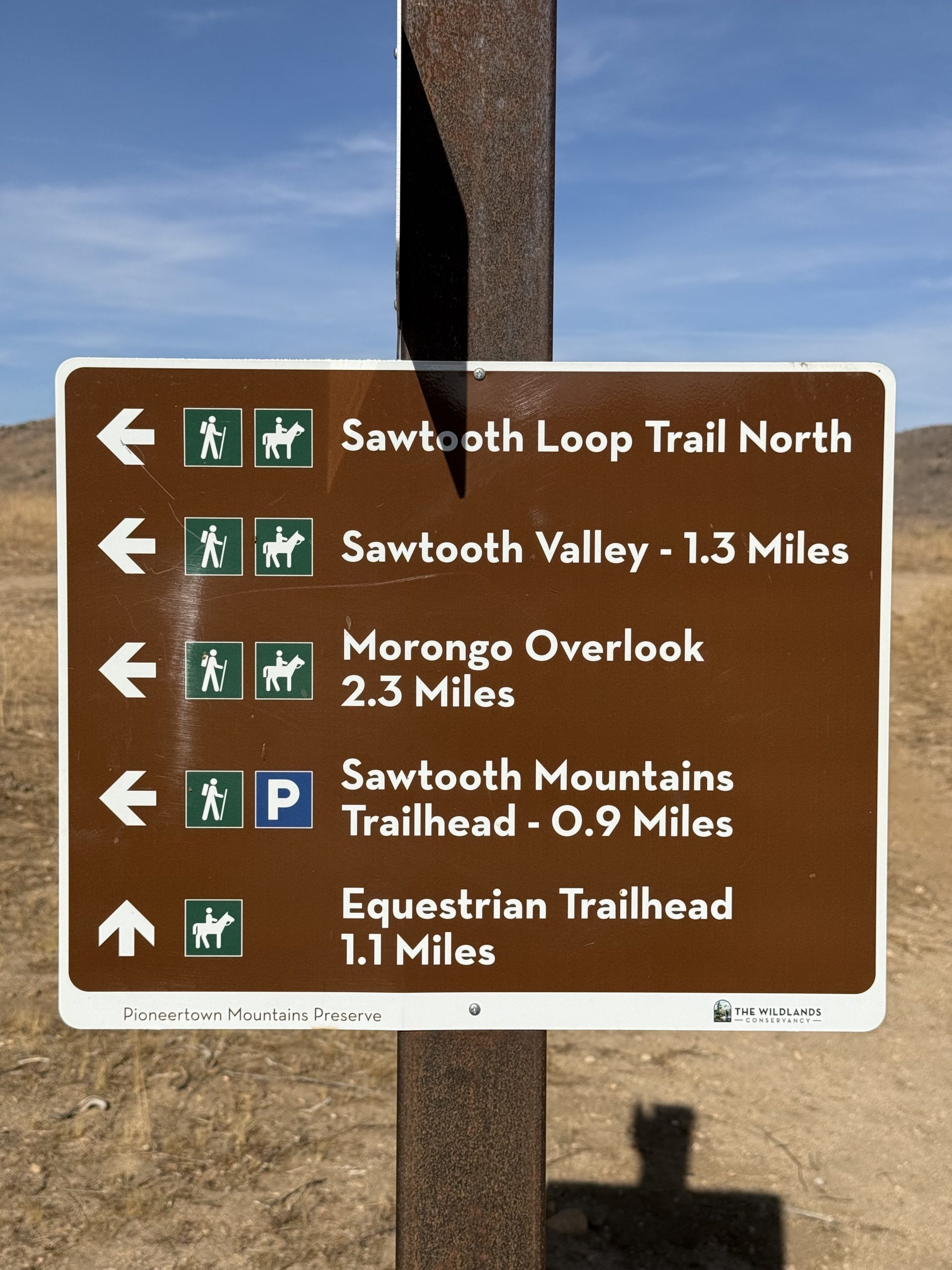 A brown outdoor sign displaying hiking trail information, including distances and directions to Sawtooth Valley, Morongo Overlook, Sawtooth Mountains Trailhead, and Equestrian Trailhead. The background features a clear blue sky and rugged terrain.