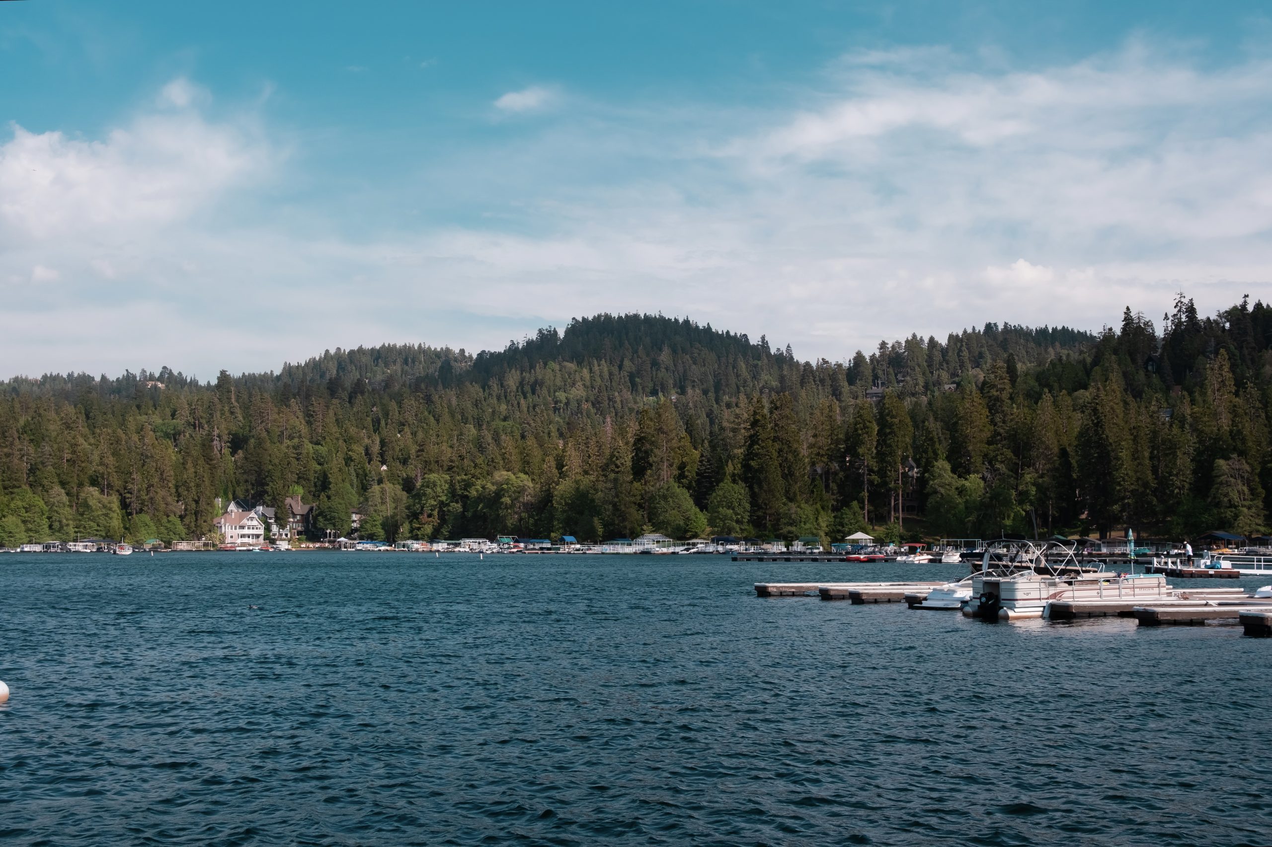 View of a peaceful lakefront with floating docks and moored boats, adjacent to a forest and a small village under a clear blue sky.